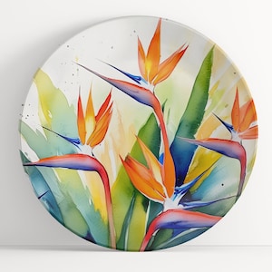 Pu&ograve; includere: Un piatto rotondo in ceramica con un dipinto ad acquerello di fiori di uccello del paradiso arancioni, blu e verdi. I fiori sono dipinti in uno stile realistico con dettagli delicati.