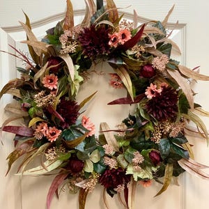 Pode incluir: Uma coroa de flores de cor bord&ocirc; e p&ecirc;ssego com folhas de eucalipto e ramos de vegeta&ccedil;&atilde;o. A coroa est&aacute; pendurada em uma porta branca com o texto "&copy;Treasured Traditions" acima.