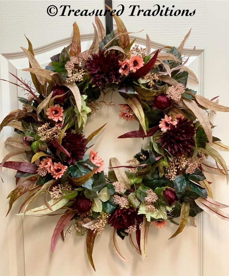 Pode incluir: Uma coroa de flores de cor bord&ocirc; e p&ecirc;ssego com folhas de eucalipto e ramos de vegeta&ccedil;&atilde;o. A coroa est&aacute; pendurada em uma porta branca com o texto "&copy;Treasured Traditions" acima.