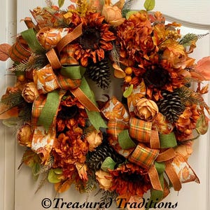 May include: A fall-themed wreath with orange, brown, and green tones. It features sunflowers, roses, pine cones, and plaid ribbon. The wreath is decorated with fall leaves and has a "Treasures Traditions" signature.
