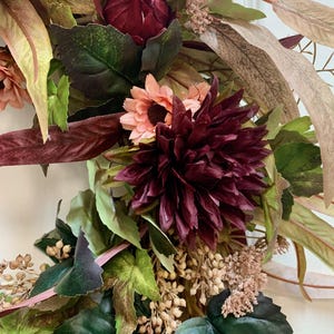 Pode incluir: Uma coroa de flores de cor bord&ocirc; e rosa com folhas verdes. A coroa &eacute; feita de flores e folhas artificiais.