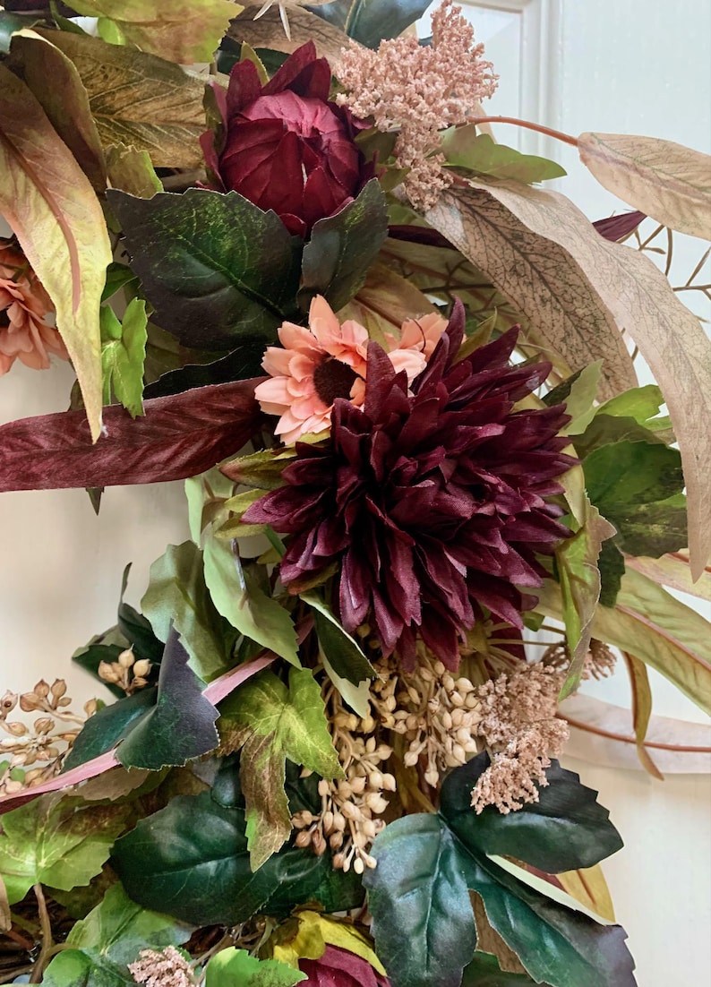 Pode incluir: Uma coroa de flores de cor bord&ocirc; e rosa com folhas verdes. A coroa &eacute; feita de flores e folhas artificiais.