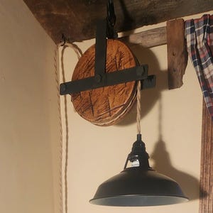 May include: An industrial-style pendant light. The black metal shade hangs from a wooden pulley system with a rope. The pulley is made of wood and black metal hardware, giving a vintage aesthetic. The light is designed for indoor use.