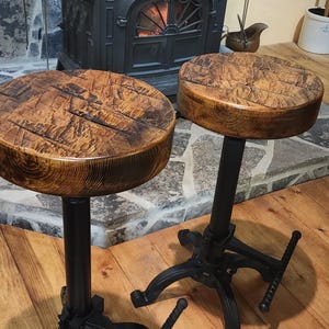 May include: Two rustic wooden bar stools with black metal bases and adjustable height levers. The stools have a distressed wood finish and are in front of a wood-burning stove.