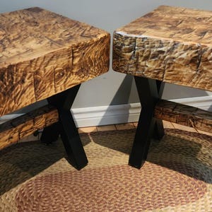 Puede incluir: Dos mesas de madera con un acabado natural y texturizado. Las encimeras rectangulares están sostenidas por patas de metal negro. Las mesas están colocadas sobre una alfombra tejida, mostrando una estética rústica. La madera tiene un tono marrón cálido.