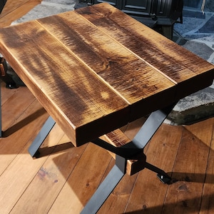Puede incluir: Una mesa de centro de madera rústica con un acabado desgastado y patas en forma de X de metal negro. La mesa está hecha de madera recuperada y tiene una veta de madera natural.
