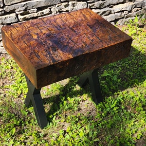 Puede incluir: Una mesa de madera rústica con un acabado marrón oscuro desgastado. La mesa tiene una tapa cuadrada y patas en forma de X de metal negro.