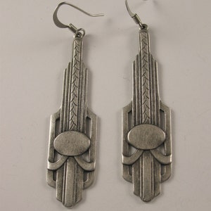 May include: A pair of silver dangle earrings with an Art Deco design. The earrings feature a geometric pattern with an oval in the center.
