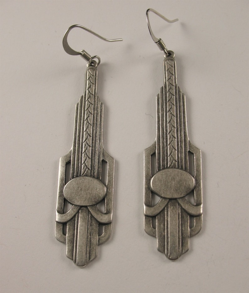 May include: A pair of silver dangle earrings with an Art Deco design. The earrings feature a geometric pattern with an oval in the center.