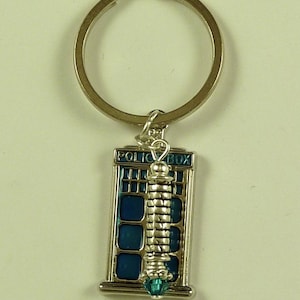 May include: A silver-toned metal keychain featuring a rectangular blue police box charm with the text POLICE BOX printed at the top. A vertical silver-toned textured column charm is attached to the front, finished with a small teal crystal bead.