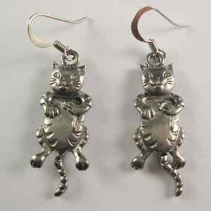 May include: A pair of silver-toned cat earrings. Each earring features a detailed, three-dimensional cat pendant with a smiling face, striped body, and curled tail. The earrings have a simple hook closure.