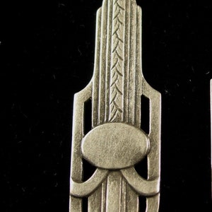 May include: Silver-toned pendant featuring an Art Deco design with geometric shapes and a central oval. The pendant has a textured surface and a detailed design.