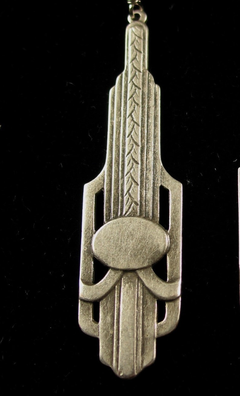 May include: Silver-toned pendant featuring an Art Deco design with geometric shapes and a central oval. The pendant has a textured surface and a detailed design.