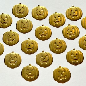 May include: A collection of nineteen small, gold-toned metal charms, each shaped like a jack-o'-lantern pumpkin with a carved face featuring triangular eyes, a triangular nose, and a jagged mouth, all arranged in a scattered pattern.