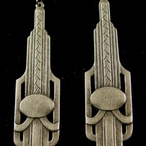 May include: A pair of silver earrings with an Art Deco design. The earrings are shaped like a tall building with a geometric pattern and a small oval in the center.