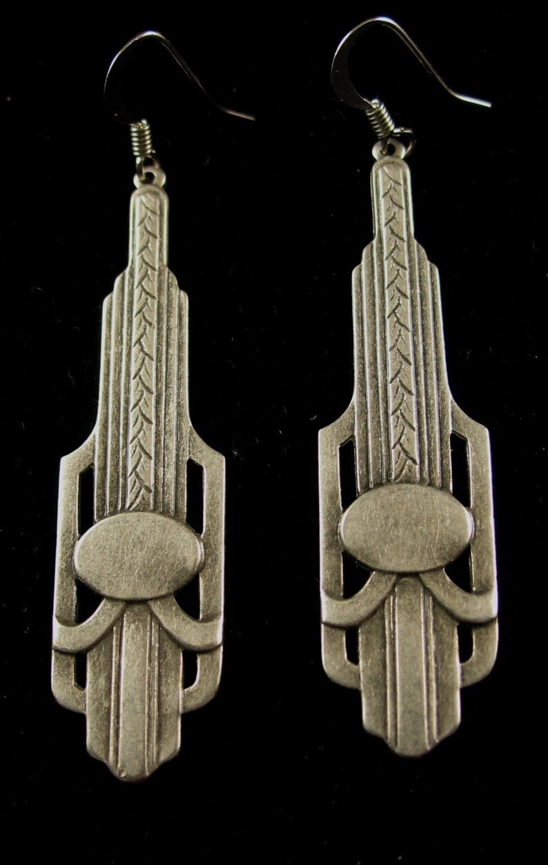 May include: A pair of silver earrings with an Art Deco design. The earrings are shaped like a tall building with a geometric pattern and a small oval in the center.