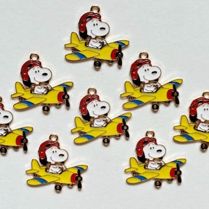 May include: A collection of ten metal charms, each featuring a white cartoon dog wearing a red aviator cap and goggles, piloting a yellow airplane with blue wing accents, a red nose, and a black propeller, with a small gold loop at the top.