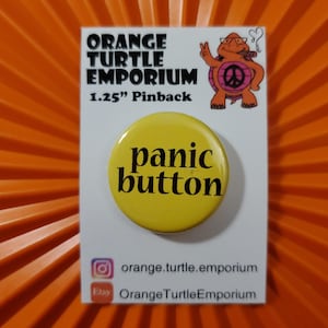 May include: A yellow 1.25 inch pinback button with the text "panic button" in black lettering. The button is on a white card with the text "Orange Turtle Emporium" in black lettering.