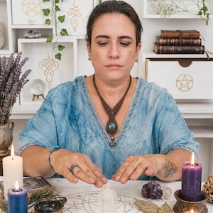 May include: A woman in a blue top with a macrame necklace, sits at a table with candles, crystals, and a clear quartz crystal. The table has a gold geometric design. Shelves in the background hold books, plants, and decorative boxes with symbols.