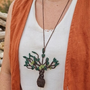 Può includere: Una collana con ciondolo ad albero fatto a mano. Il ciondolo presenta un design dettagliato ad albero con foglie verdi e un tronco marrone, sospeso a un cordino marrone. L'albero ha un piccolo cristallo trasparente in basso. La persona indossa un top bianco e un gilet arancione.