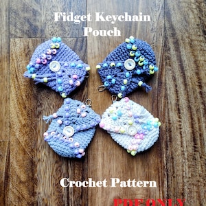 May include: Four handmade crochet fidget keychain pouches in shades of blue and white, adorned with colorful beads and button closures. The text "Fidget Keychain Pouch" and "Crochet Pattern" are displayed on a wooden surface.