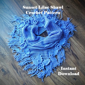 Puede incluir: Un chal de ganchillo azul con flecos y detalles florales. El texto "Sunset Lilac Shawl Crochet Pattern" y "Instant Download" son visibles. El chal se muestra sobre una superficie de madera.