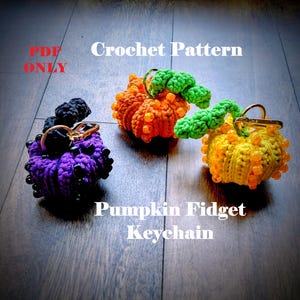 May include: Three crochet pumpkin keychains in orange, yellow, and purple. Each pumpkin has a green stem and is adorned with beads. The keychains are labelled "Crochet Pattern" and "Pumpkin Fidget Keychain."