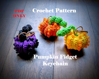 Op de afbeelding: Drie gehaakte pompoen sleutelhangers in oranje, geel en paars. Elke pompoen heeft een groene stengel en is versierd met kralen. De sleutelhangers zijn voorzien van de tekst "Crochet Pattern" en "Pumpkin Fidget Keychain."
