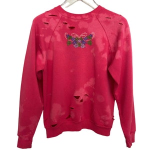 Könnte beinhalten: Rosafarbener Pullover mit ausgefransten Löchern und einem floralen Stickmuster auf der Rückseite.