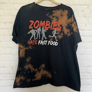 Könnte beinhalten: Schwarzes T-Shirt mit einem Design aus Bleichmittelspritzern. Das Shirt zeigt eine Grafik von Zombies mit dem Text "Zombies Hate Fast Food".