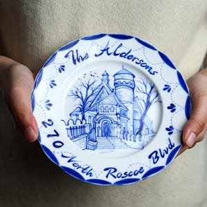 May include: A white ceramic plate with a blue hand-painted illustration of a building and text. The plate has a scalloped edge with blue accents and the words "The Aldersons" and "270 North Roscoe Blvd".