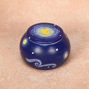 May include: A small, round, navy blue ceramic jar with a lid. The lid and sides are painted with a starry night design, featuring a yellow crescent moon, white stars, and swirling lines. The jar is a decorative item.