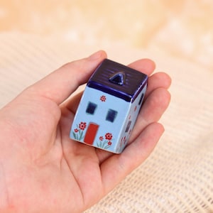 May include: A small, ceramic house figurine with a light blue body and a dark blue roof. The house features a red door, dark blue windows, and red flower accents. The figurine is held in a hand.