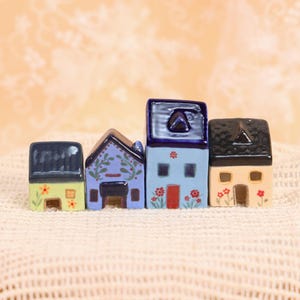 May include: Four miniature ceramic houses in a row. Each house has a unique design, with varying colors including blue, yellow, and beige. Floral accents and detailed roof designs add charm to the tiny dwellings.