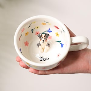 K&ouml;nnte beinhalten: Wei&szlig;e Keramiktasse mit einer 3D-Hundefigur im Inneren, umgeben von bunten Blumenmustern. Die Tasse hat einen Henkel und der Name "Skye" steht auf dem Boden. Die Tasse wird von einer Person gehalten.