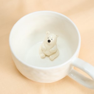 May include: White ceramic mug featuring a polar bear figurine. The bear is seated inside the mug, with a black nose and eyes. The mug has a curved handle and a smooth, glossy finish. The mug is ideal for hot beverages.