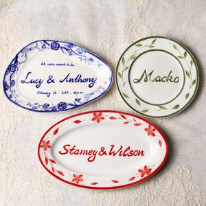 May include: Three decorative ceramic platters with handwritten text. One blue platter reads "Lucy & Anthony" with a date. Another reads "Macko" with green leaves. The third reads "Stamey & Wilson" with red flowers.