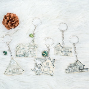 May include: Six different house-shaped keychains with silver key rings and chains. Each keychain has a different design of a house with black and white details. Some keychains have a green and blue bead charm.