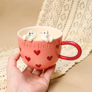 May include: Pink ceramic mug with red heart designs and the text "Mix & Nala". The mug features a red handle and is decorated with white dog and cat figurines on the rim.