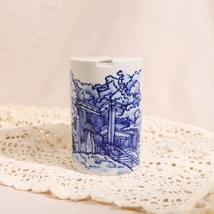 Pode incluir: Um recipiente cilíndrico de cerâmica branca com uma ilustração detalhada em azul de estilo transferência, apresentando uma casa de vários andares com alpendre, escadas e árvores. O topo do recipiente tem uma pequena abertura retangular.