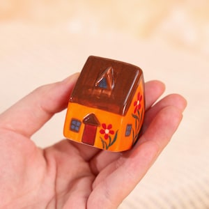 May include: A small, hand-painted ceramic house figurine. The house features an orange body, a brown roof, and red and blue accents. Decorative red flowers adorn the side. The figurine is held in a hand.