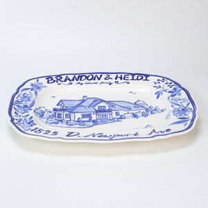 May include: A white ceramic serving platter with a blue floral border and a blue illustration of a house. The platter has the text "BRANDON & HEIDI My beloved family" and "1825 D. Newport Ave".