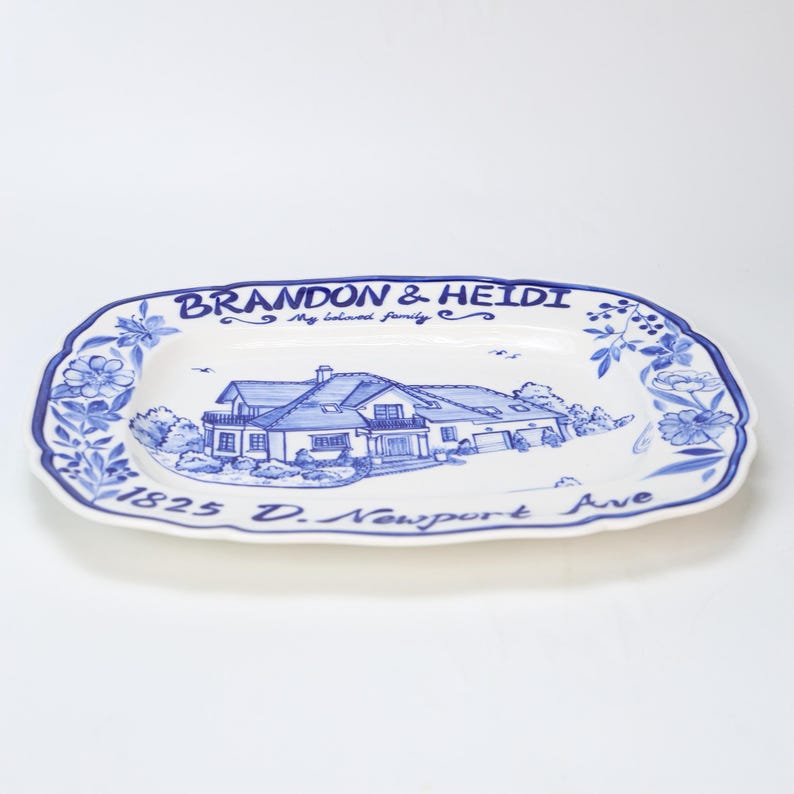 May include: A white ceramic serving platter with a blue floral border and a blue illustration of a house. The platter has the text "BRANDON & HEIDI My beloved family" and "1825 D. Newport Ave".