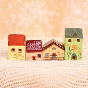 Könnte beinhalten: Vier Miniatur-Keramikhäuser in einer Reihe. Jedes Haus hat ein einzigartiges Design mit roten und grünen Dächern. Die Häuser sind in Gelb, Pfirsich und Grün bemalt und weisen Blumen- und Fensterdetails auf.