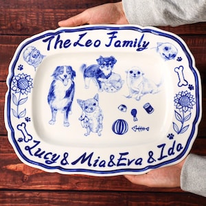 May include: A white ceramic serving platter with a blue scalloped edge, decorated with blue illustrations of dogs and dog-themed items. The text "The Leo Family" and "Lucy & Mia & Eva & Ido" are written in blue.
