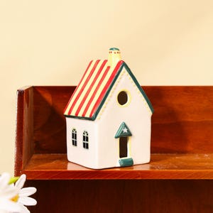 Könnte beinhalten: Eine Keramikhausfigur mit einem rot-gelb gestreiften Dach, grünen Zierleisten und einem Schornstein. Das Haus hat zwei kleine Fenster und eine Tür mit einem Stern darüber. Das Haus ist weiß mit einem runden Loch in der Nähe des Daches.
