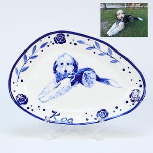 Könnte beinhalten: Weiße Keramikplatte mit einer blau-weißen Hundeillustration. Die Platte hat einen blauen Blumenrand mit Rosen- und Blättern. Der Hund trägt ein blaues Halstuch. Der Name "Koa" ist auf der Platte geschrieben.