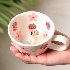 Pode incluir: Uma caneca de cerâmica branca com uma alça, decorada com flores cor-de-rosa e morangos vermelhos. Dentro da caneca há uma pequena figura de um cão castanho e branco.