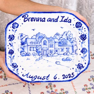 Peut inclure: Un plat en céramique blanc et bleu décoratif avec une illustration peinte à la main d'une maison et d'un jardin. Le plat est bordé de roses bleues et porte les noms "Brenna and Ida" et la date "6 août 2025".