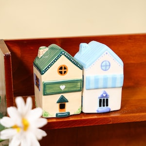 Pode incluir: Duas figuras de casas de cerâmica. Uma casa é bege com telhado e acabamentos verdes, um coração e uma janela laranja. A outra casa é branca com telhado e acabamentos azuis e uma porta roxa.